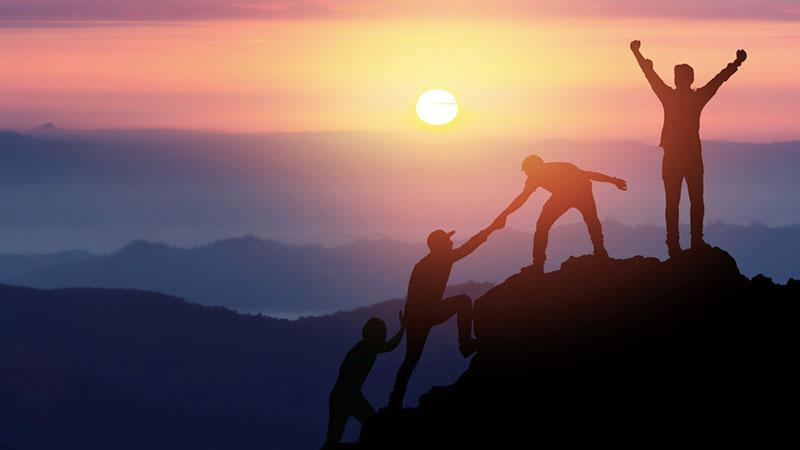 Silhouettes of people helping each other climb a mountain at sunrise.