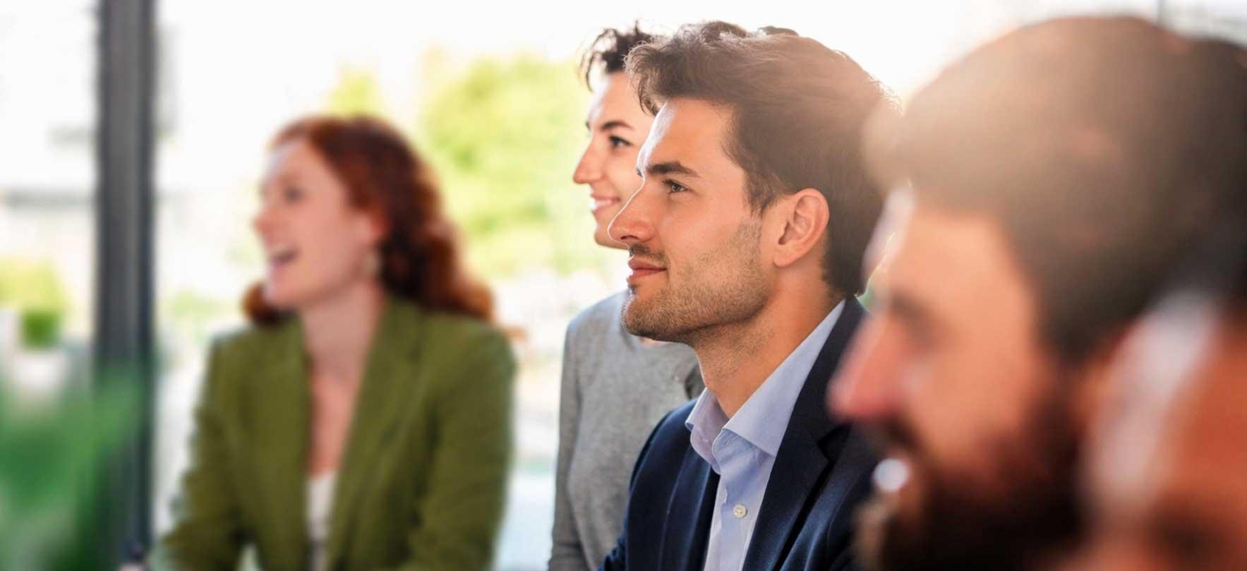 Male student in a business school classroom, listening attentively, with other students blurred in the background.