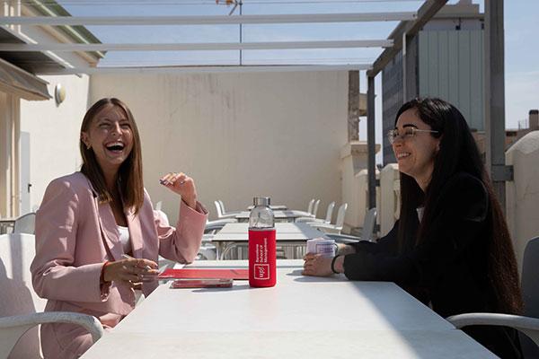 Two students laughing and chatting at an outdoor terrace table on a sunny day, with UPF-BSM water bottle on the table.