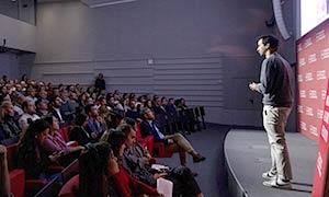 Speaker giving a presentation to a full audience in an auditorium at UPF-BSM, with a branded red backdrop on stage.